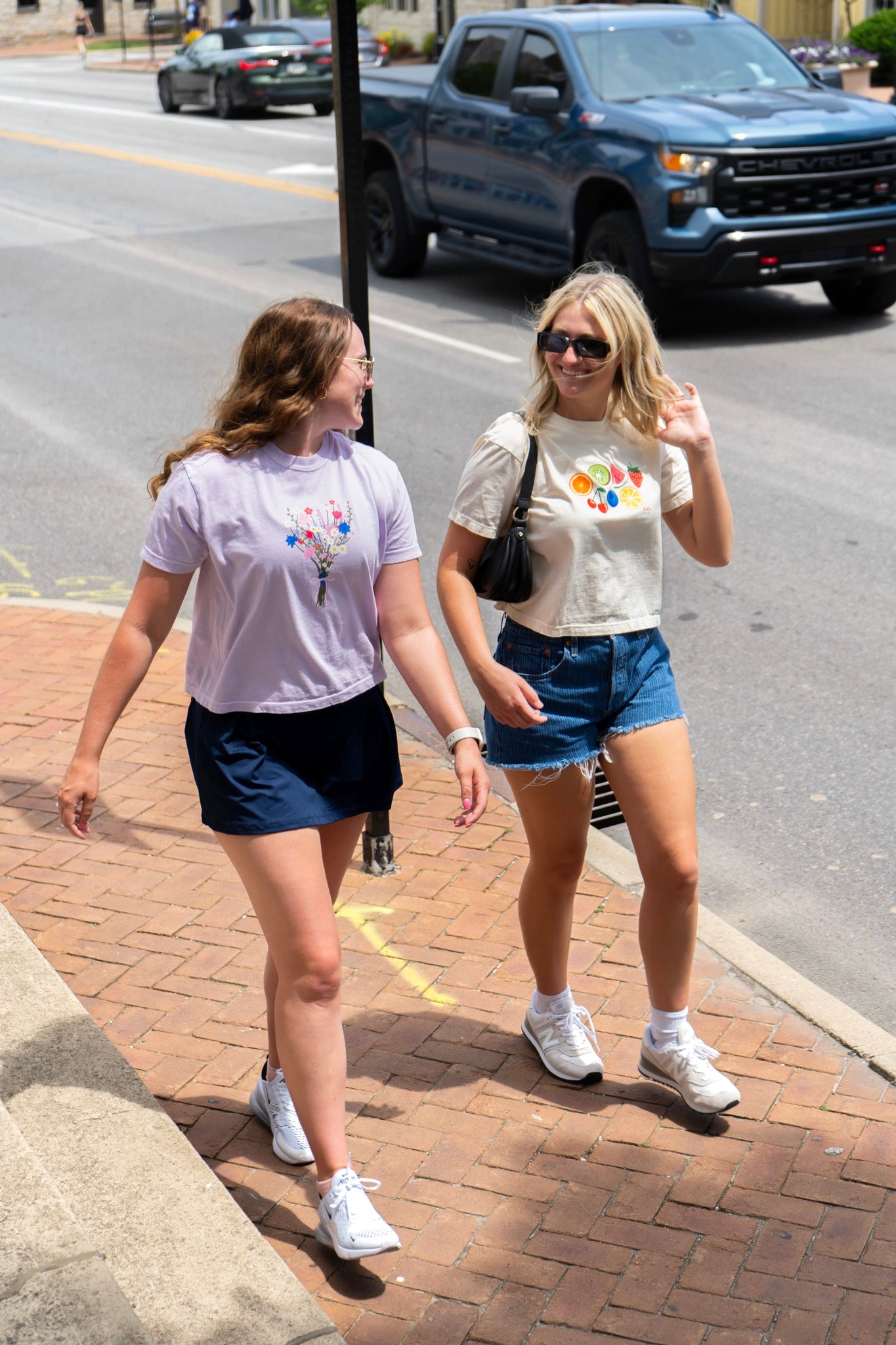 Two women walking on a sidewalk wearing the fruit tshirt and wildflower tshirt with a street and cars in the background